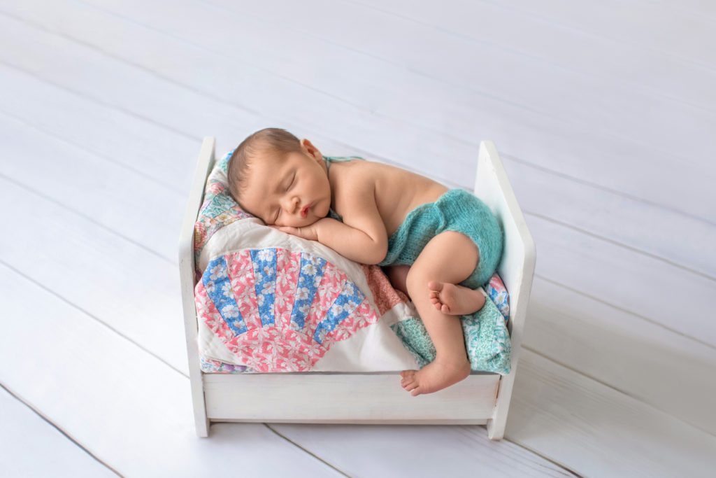 Newborn boy Jeffery outfitted in aqua romper asleep with chin resting on his hand lying on his side on charming quilt covered white wooden bed newborn photographer Gainesville Florida