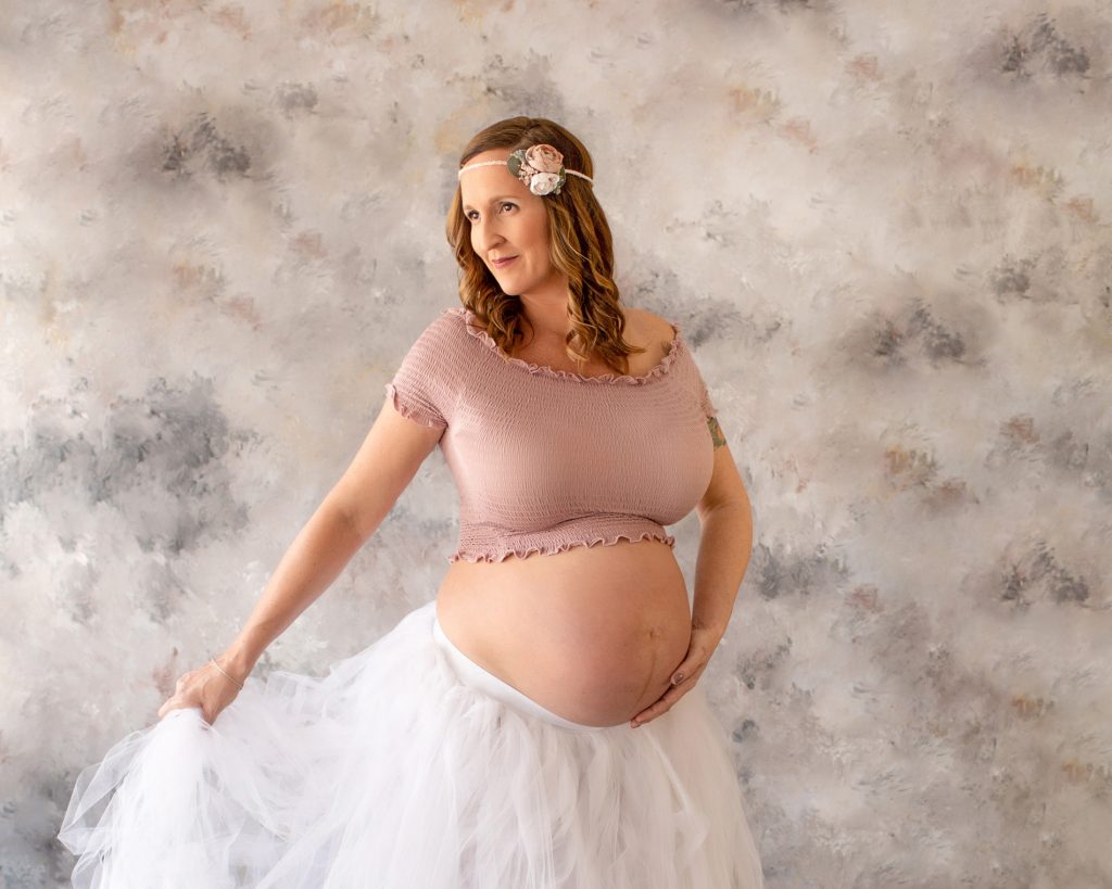 Beautiful pregnant mom to be in white tulle tutu posed with hand on bare belly and dreamy eyes against pastel blue grey rose painted backdrop Gainesville maternity photography