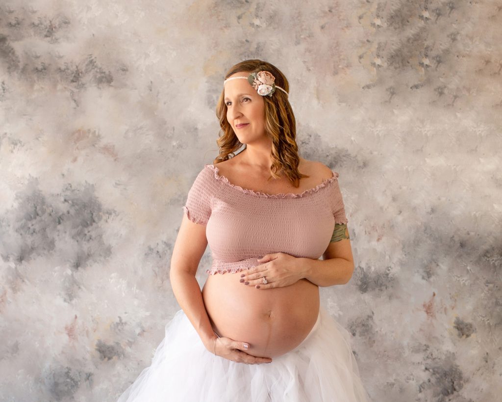 Beautiful pregnant mom to be with eyes of anticipation dressed in white tulle tutu posed with hands on bare belly against pastel blue grey rose painted backdrop Gainesville maternity photography