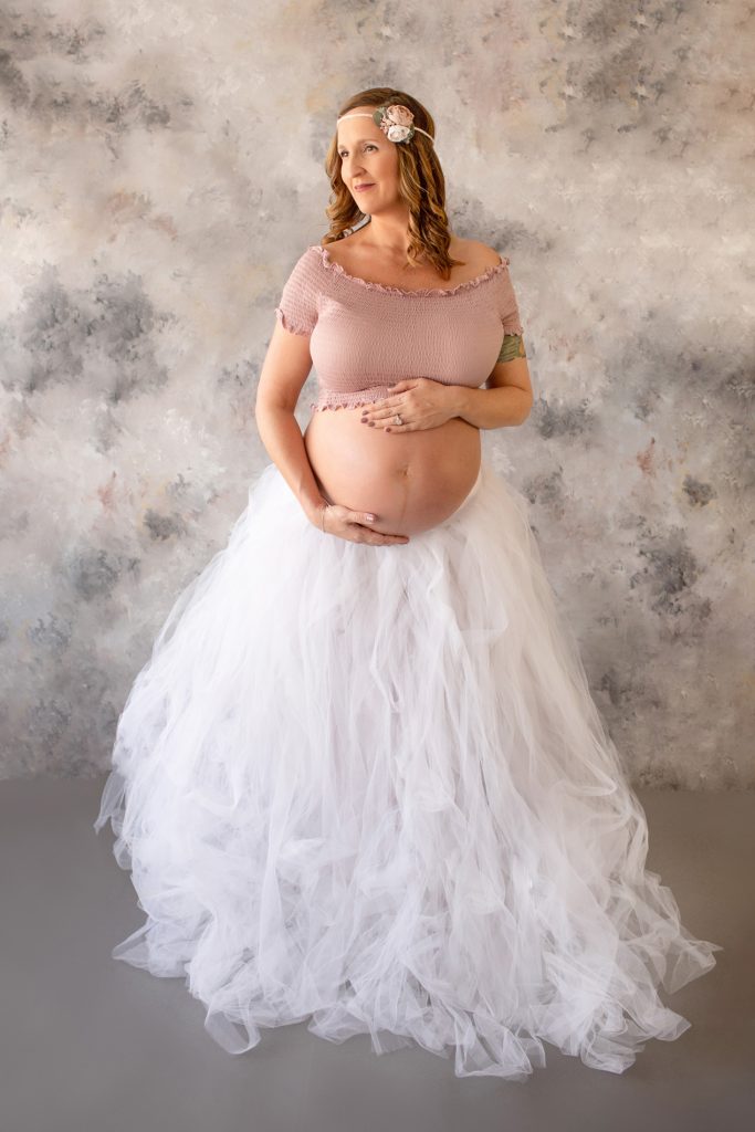 Beautiful pregnant mom to be in white tulle tutu posed with hands on bare belly against pastel blue grey rose painted backdrop Gainesville maternity photography
