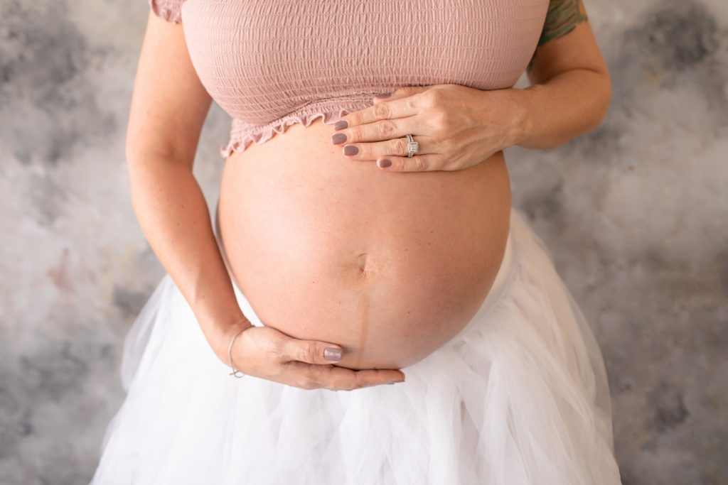 Beautiful pregnant mom to be in white tulle tutu posed with close up hands on bare belly against pastel blue grey rose painted backdrop Gainesville maternity photography