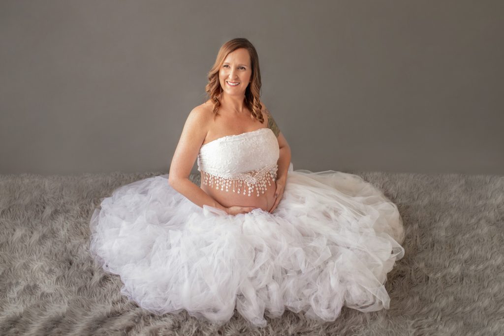 Beautiful pregnant mom to be in white tulle tutu and beaded white lace top posed on grey fur
