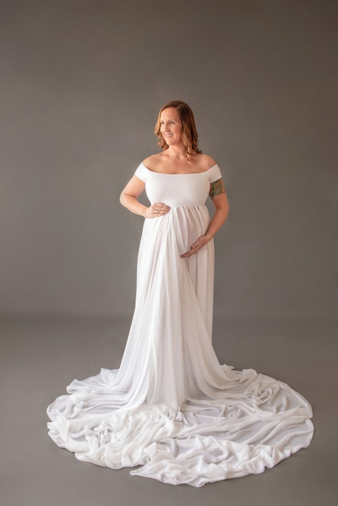 Beautiful pregnant mom in white chiffon maternity gown holding belly and looking sideways on grey background