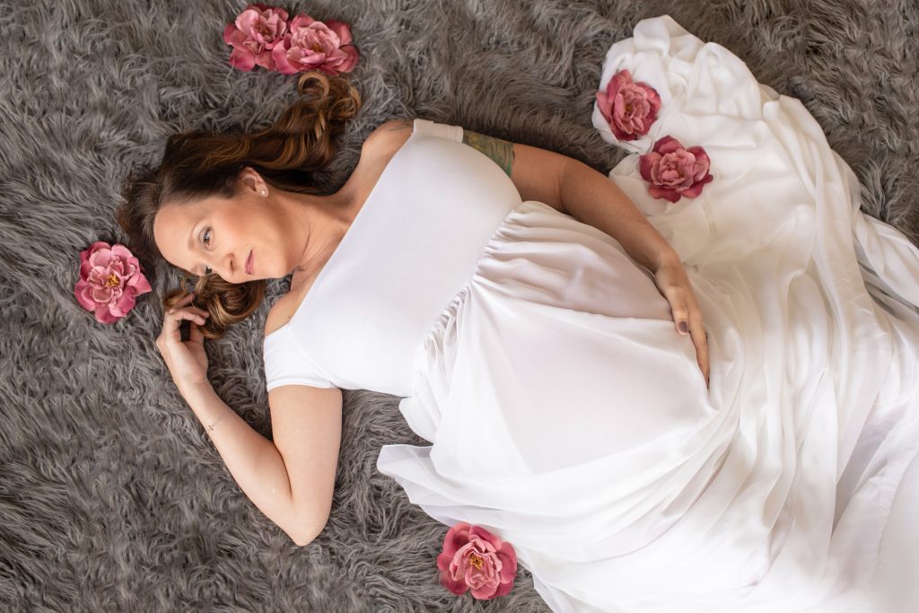Beautiful pregnant mom to be in white chiffon maternity gown lying on grey fur with dusty rose flowers Gainesville maternity photography