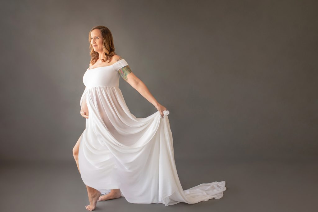 Beautiful pregnant mom in white chiffon maternity gown opened for belly to poke through with look of anticipation on grey background