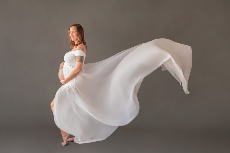 Gorgeous maternity photo pregnant mom in white chiffon maternity gown opened for belly to poke through with gown toss and wind blown train gown on grey background Gainesville maternity photography