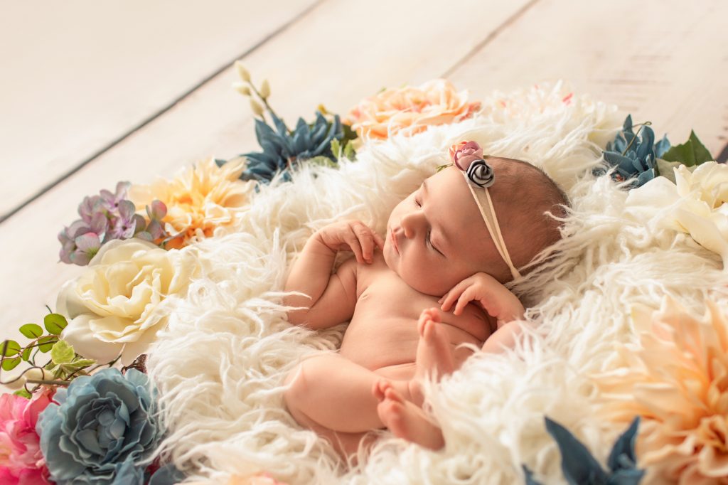 Gainesville Newborn moments newborn baby girl posed naked with delicate floral headband against white fur surrounded by dusty blue pink and ivory flowers photo with backlight Gainesville Florida newborn photography