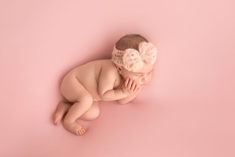 Gainesville Newborn moments baby girl posed naked with pink handmade knit headband newborn baby hands cupping her cheeks posed on her side on dust pink blanket photo from above Gainesville Florida newborn photography