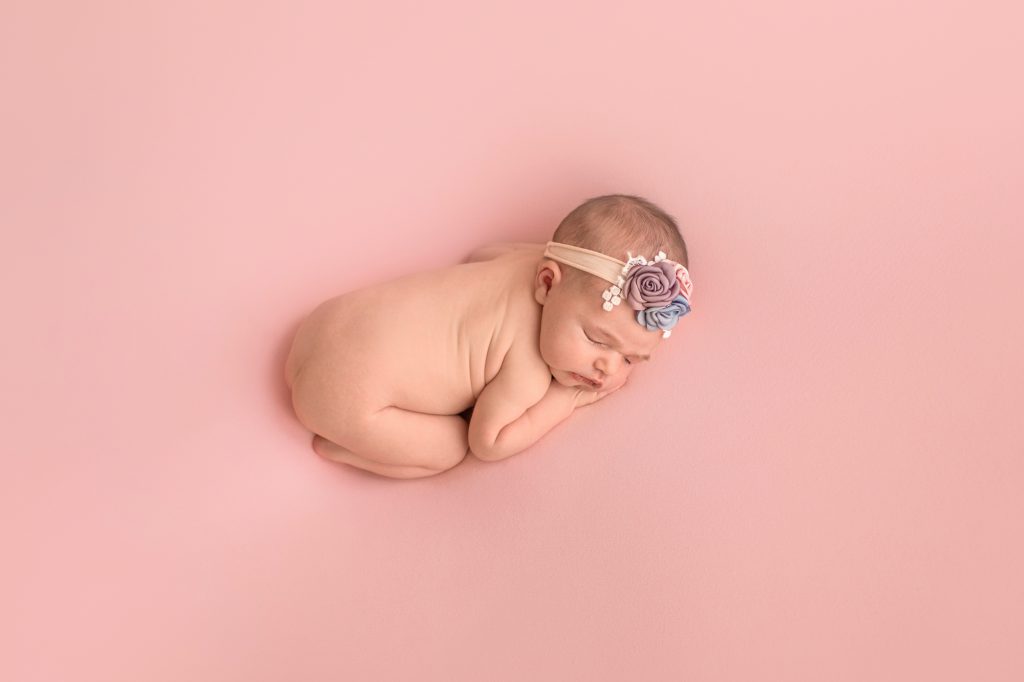 Gainesville Newborn moments newborn baby girl posed naked with dusty blue pink lavender floral headband newborn baby posed on belly with bottom up resting head on her hands lying on dusty pink blanket photo from above Gainesville Florida newborn photography