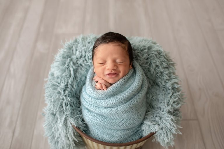 newborn photoshoot smiles big from ear to ear in aqua knit blanket wrapped like potato sack with newborn hands folded below chin posed in aqua fur stuffed bucket on grey wood floor