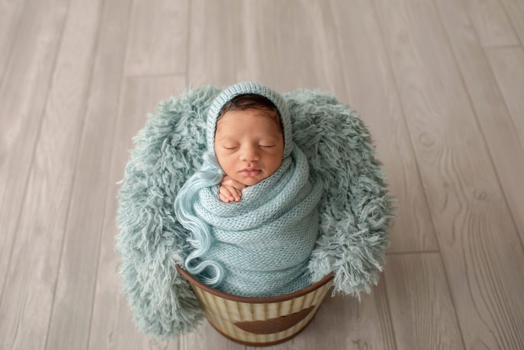 newborn boy Christian in aqua knit blanket wrapped like potato sack with newborn hands folded below chin matching aqua knit bonnet posed in aqua fur stuffed bucket facing forward on grey wood floor