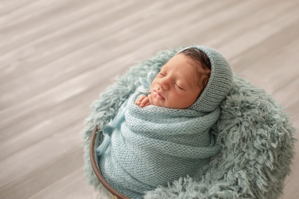 newborn boy Christian in aqua knit blanket wrapped like potato sack with newborn hands folded below chin matching aqua knit bonnet posed in aqua fur stuffed bucket on grey wood floor photo from side