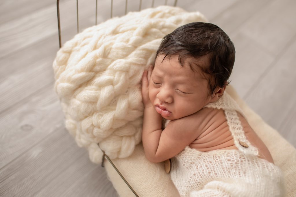 close up newborn photoshoot boy Christian wears cream knit romper with buttons posed with hand under cheek on cream covered metal bed Gainesville Fl newborn picture