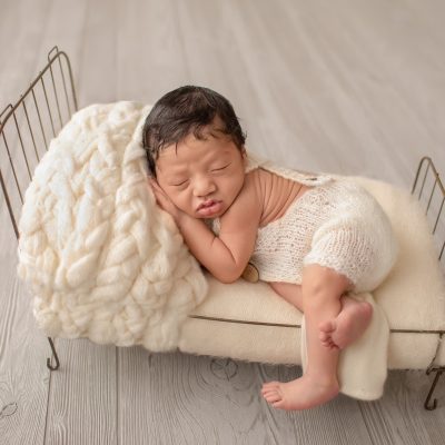 newborn wears cream knit romper with buttons posed with hand under cheek on cream covered metal bed with leg hanging off side of bed