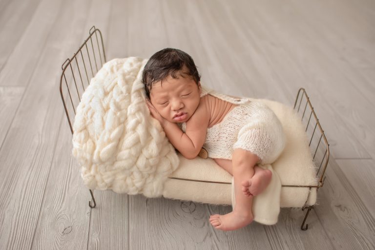 newborn wears cream knit romper with buttons posed with hand under cheek on cream covered metal bed with leg hanging off side of bed