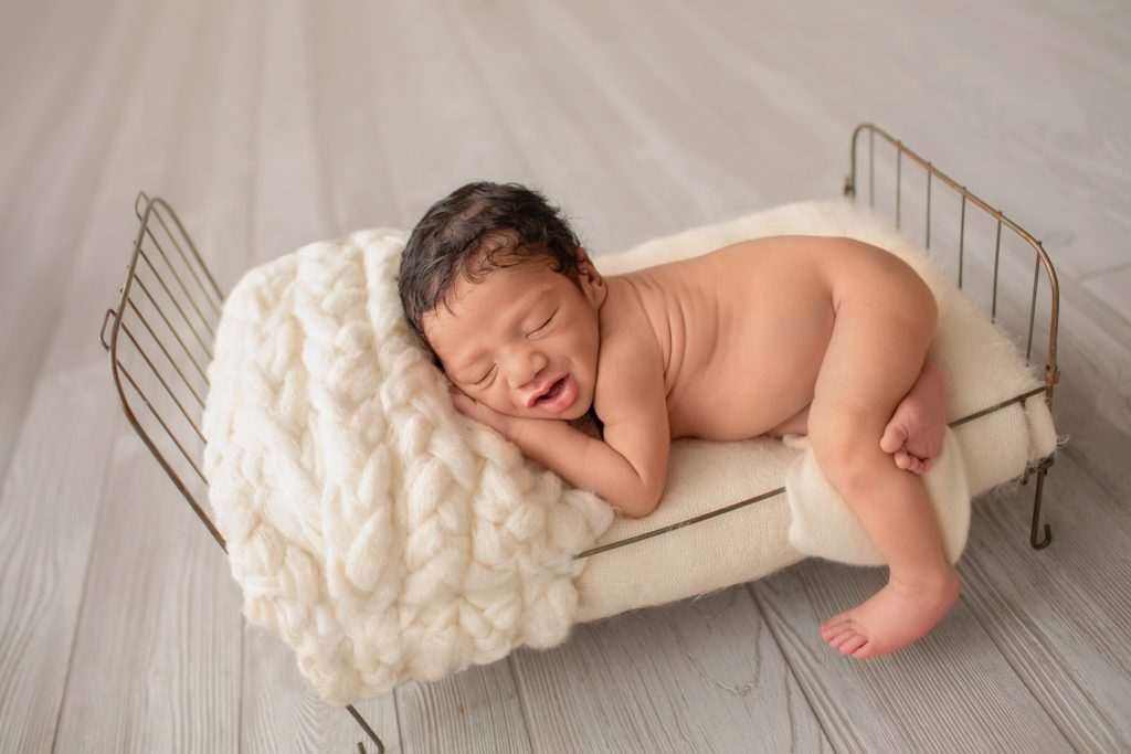 newborn photoshoot boy Christian smiling posed naked with hand under cheek on cream covered metal bed with leg hanging off side of bed Gainesville Fl newborn picture