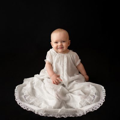 6 month old Rachel family heirloom photos smiling posed in 205 year old baby christening gown sitting up against a solid black backdrop