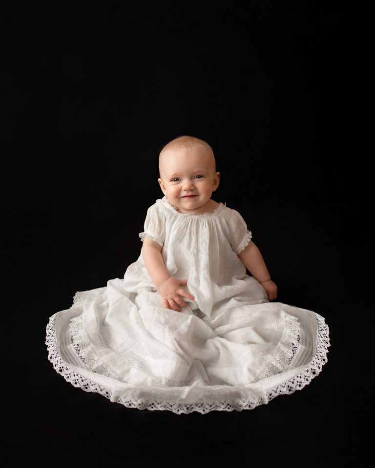 6 month old Rachel family heirloom photos smiling posed in 205 year old baby christening gown sitting up against a solid black backdrop