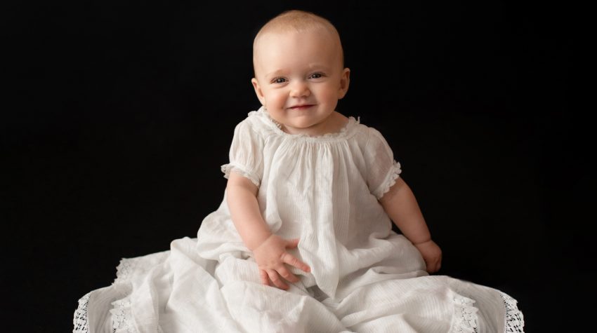 6 month old Rachel family heirloom photos smiling posed in 205 year old baby christening gown sitting up against a solid black backdrop