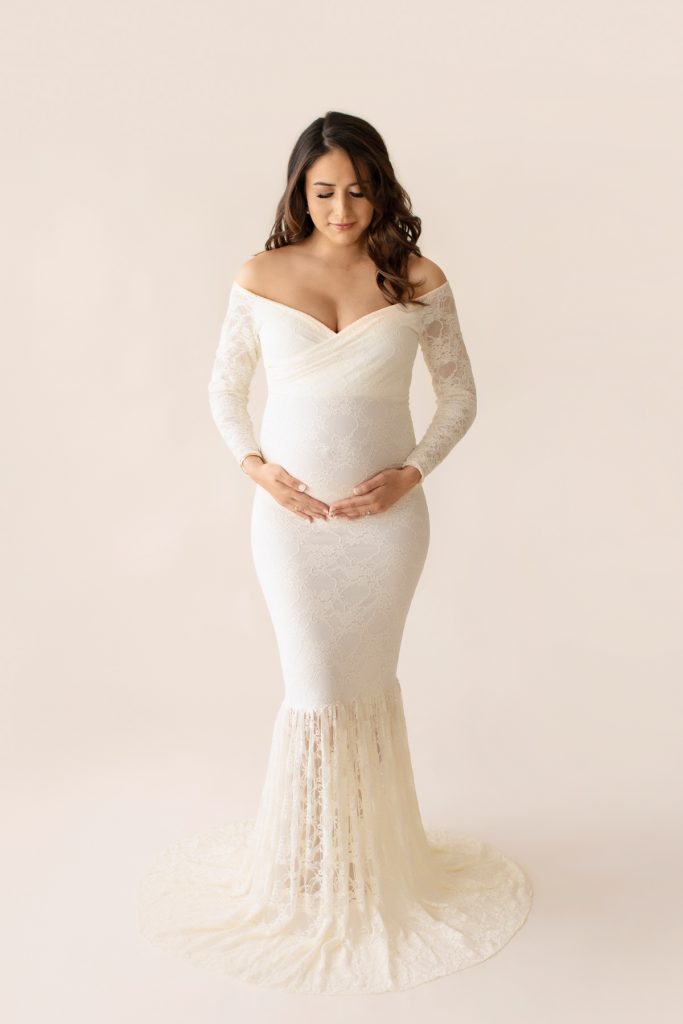 Gorgeous pregnant mom dressed in long sleeved ivory lace mermaid maternity gown looking down at beautiful baby belly