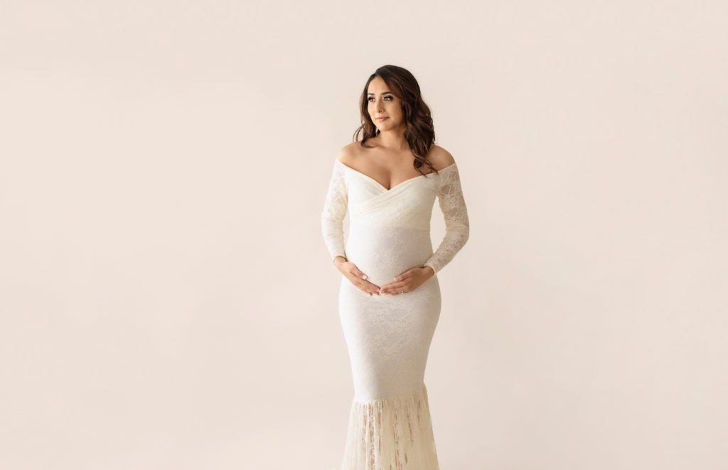Gorgeous pregnant mom dressed in long sleeved ivory lace mermaid maternity gown looking off to the side and dreaming about baby maternity pictures