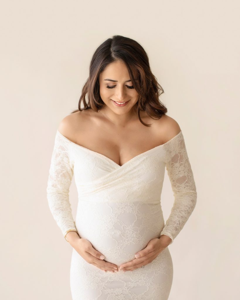 Gorgeous pregnant mom dressed in long sleeved ivory lace mermaid maternity gown smiling looking down at beautiful baby belly