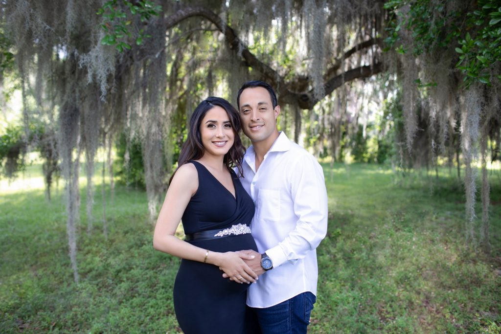 Beautiful pregnant mom dressed in black sleeveless v-neck maternity gown posed with dad at lush green park