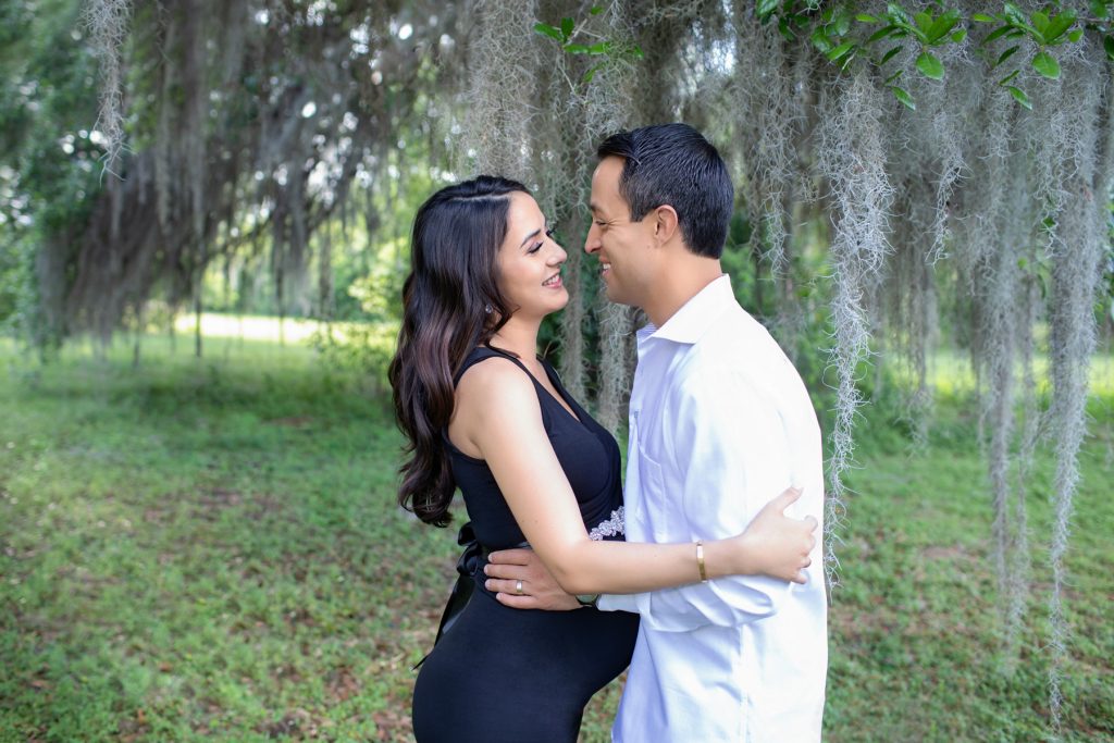 Beautiful pregnant mom dressed in black sleeveless v-neck maternity gown laughing posed profile facing dad at lush green park