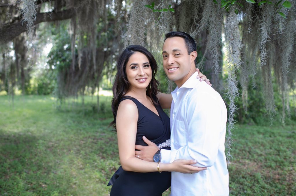 Beautiful pregnant mom dressed in black sleeveless v-neck maternity gown posed profile facing dad at lush green park