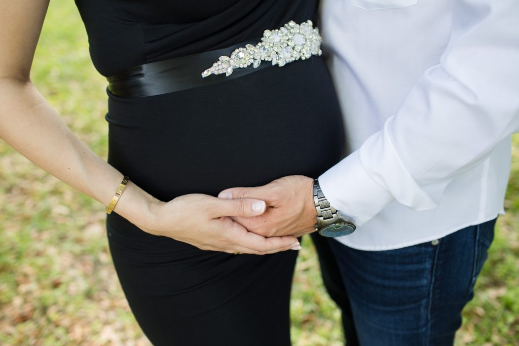 Beautiful pregnant mom dressed in black sleeveless v-neck maternity gown posed holding beautiful baby belly with dad close up at lush green park