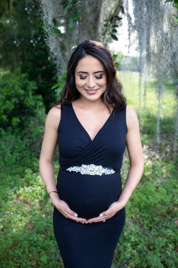 Beautiful pregnant mom dressed in black sleeveless v-neck maternity gown looking down at beautiful baby belly at lush green park