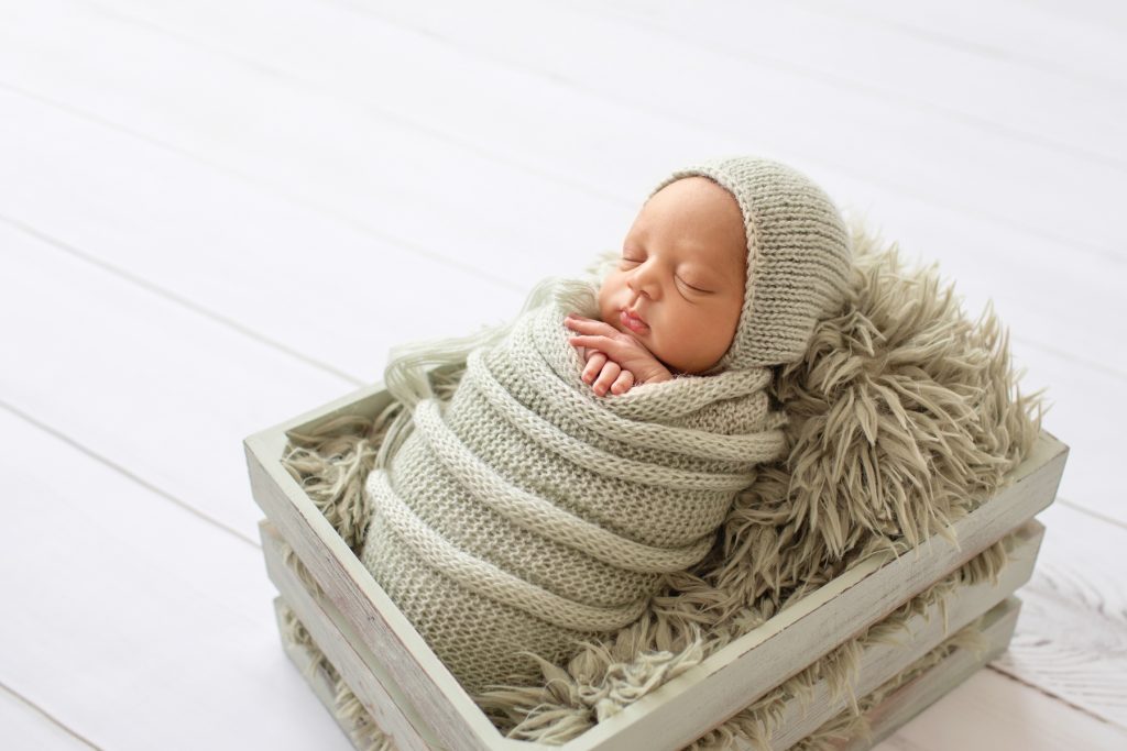 Baby boy snuggled with sage green knit wrap and bonnet on sage fur in crate