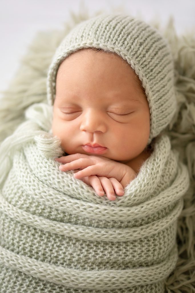 Baby boy close up chubby cheeks folded baby hands snuggled with sage green knit wrap and bonnet on sage fur in crate