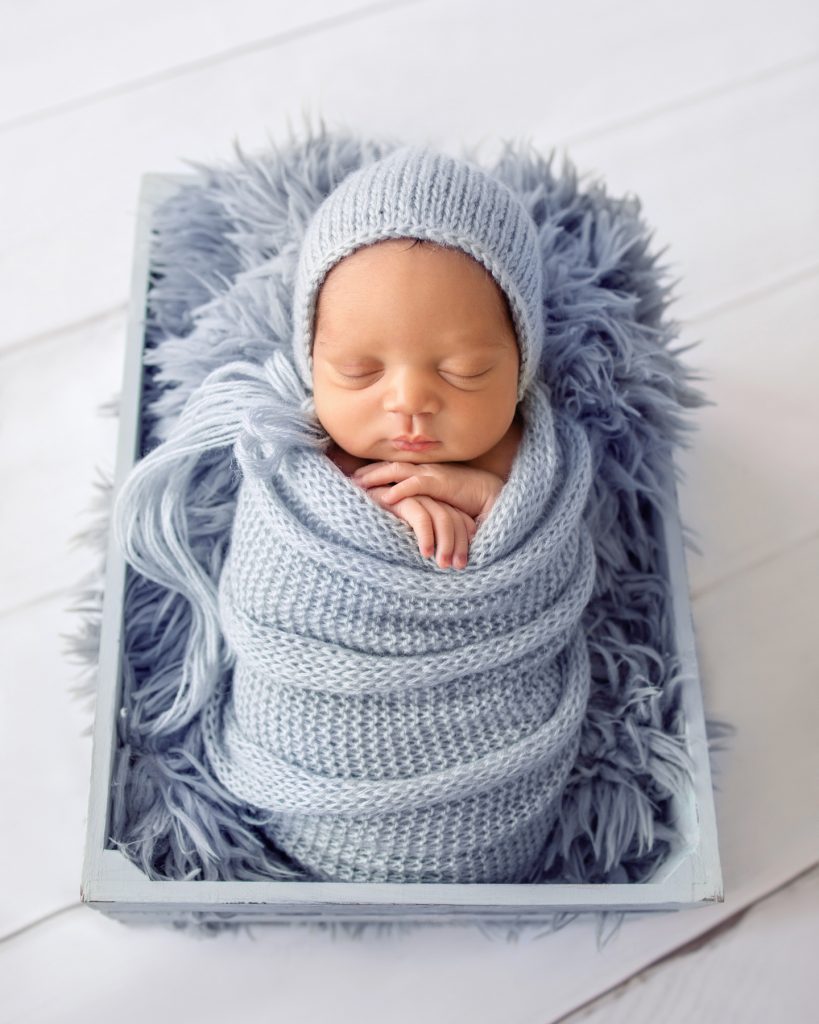 Baby boy chubby cheeks folded baby hands snuggled with light blue knit wrap and bonnet on blue fur in crate