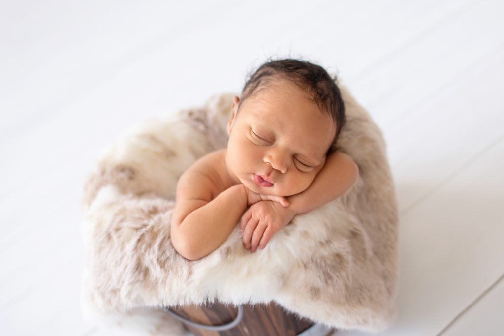 Baby boy chubby cheeks leans on his folded baby hands on rabbit fur in brown bucket