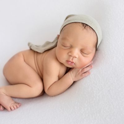 newborn baby photos naked boy chubby cheeks sleeps posing on his side with head resting on hand beautiful baby skin wearing a sage sleepy hat on white blanket