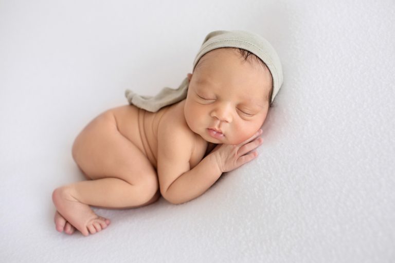 newborn baby photos naked boy chubby cheeks sleeps posing on his side with head resting on hand beautiful baby skin wearing a sage sleepy hat on white blanket