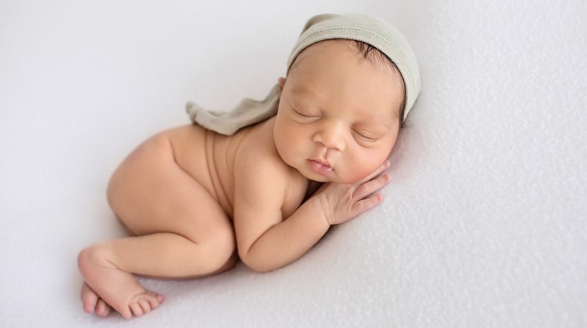 newborn baby photos naked boy chubby cheeks sleeps posing on his side with head resting on hand beautiful baby skin wearing a sage sleepy hat on white blanket