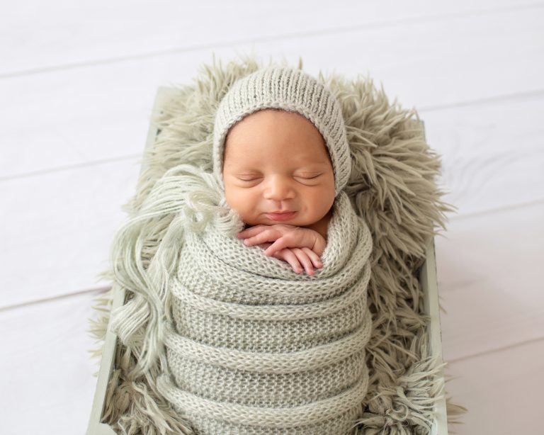 newborn baby photos boy with big smile and chubby cheeks folded baby hands snuggled with sage green handknit wrap and bonnet on sage fur in crate