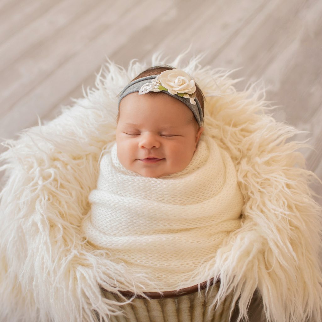 newborn baby girl smiles Bryce swaddlied in white hand knitted blanket posed like a little potato sack in a bucket with white fur