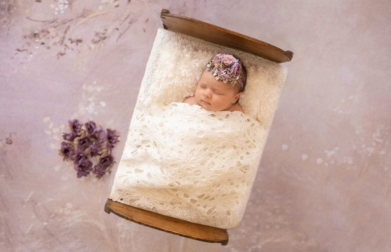 tiny baby girl Bryce sleeps under white lace blanket in baby bed with purple flowers and matching headband
