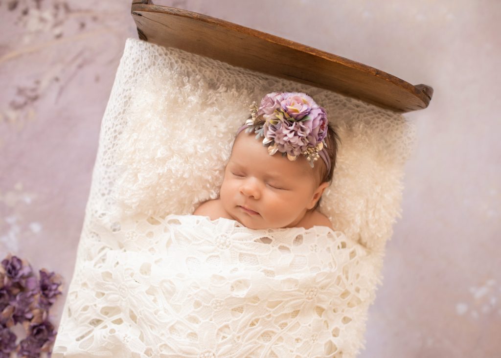 Close up tiny baby girl Bryce sleeps under white lace blanket in baby bed with purple flowers and matching headband