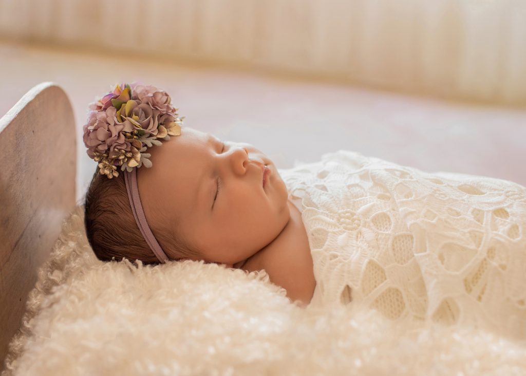 Profile tiny baby girl Bryce sleeps under white lace blanket in baby bed with purple flowers and matching headband