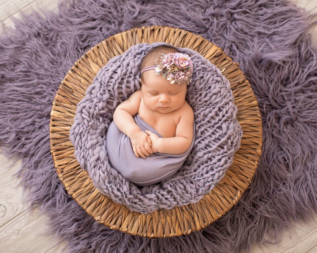 Newborn girl Bryce holds her hands with purple wrap and fur in brown basket