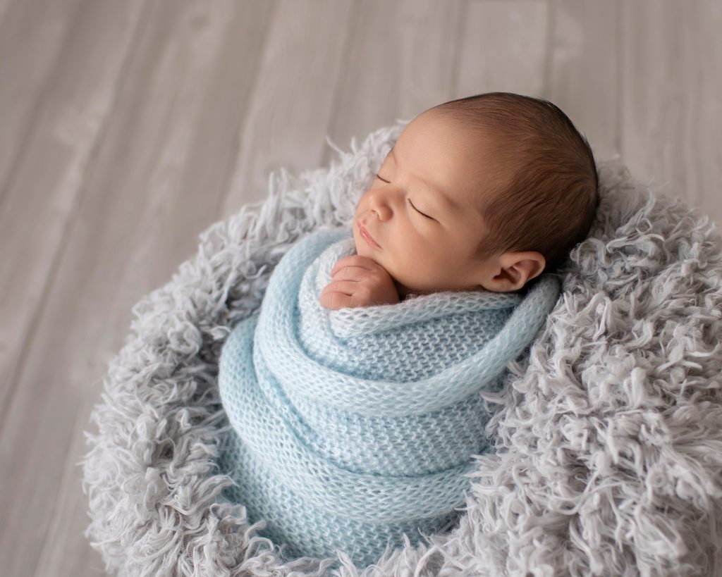 baby from the side in grey stuffed bowl wrapped in pale blue knit blanket against grey floor
