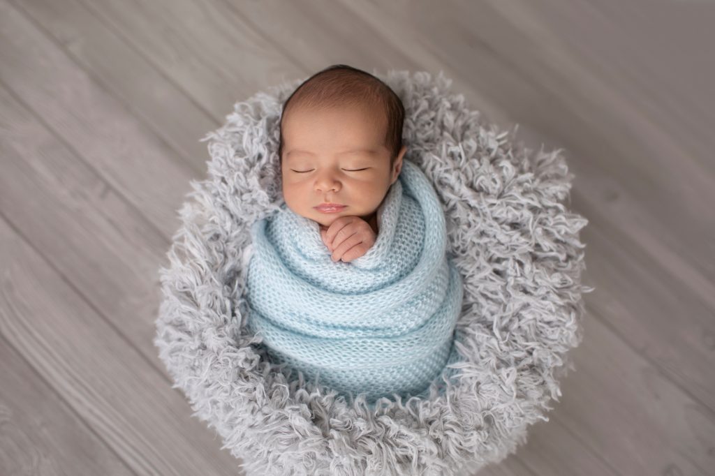 baby in grey stuffed bowl wrapped in pale blue knit blanket against grey wood floor