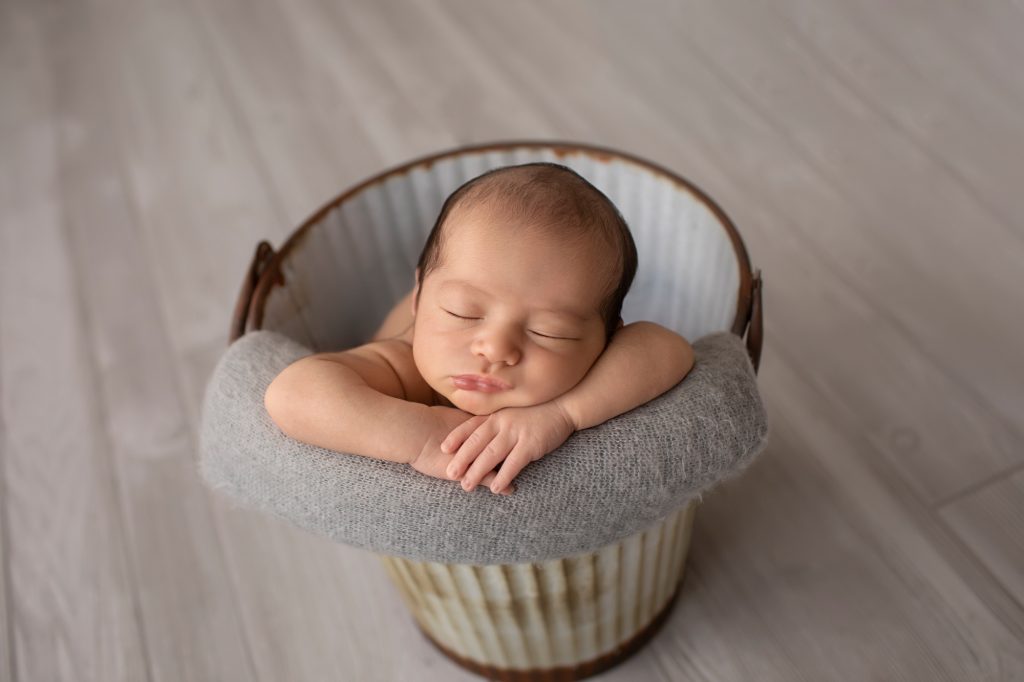 newborn image naked in metal bucket against grey wood floor