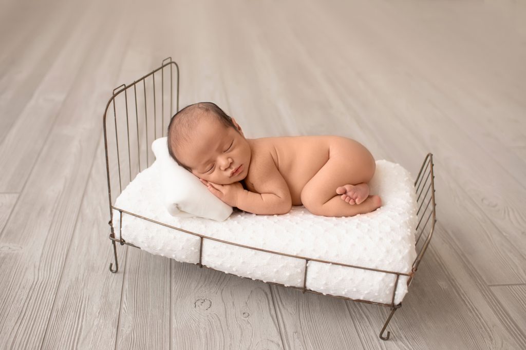 newborn image naked posed on metal bed bottom up hands under face white dotted bedspread grey wood floor