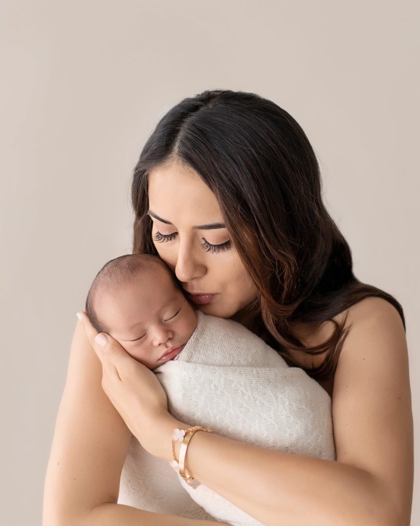 Mom kissing newborn bundled in cream knit blanket cuddle time