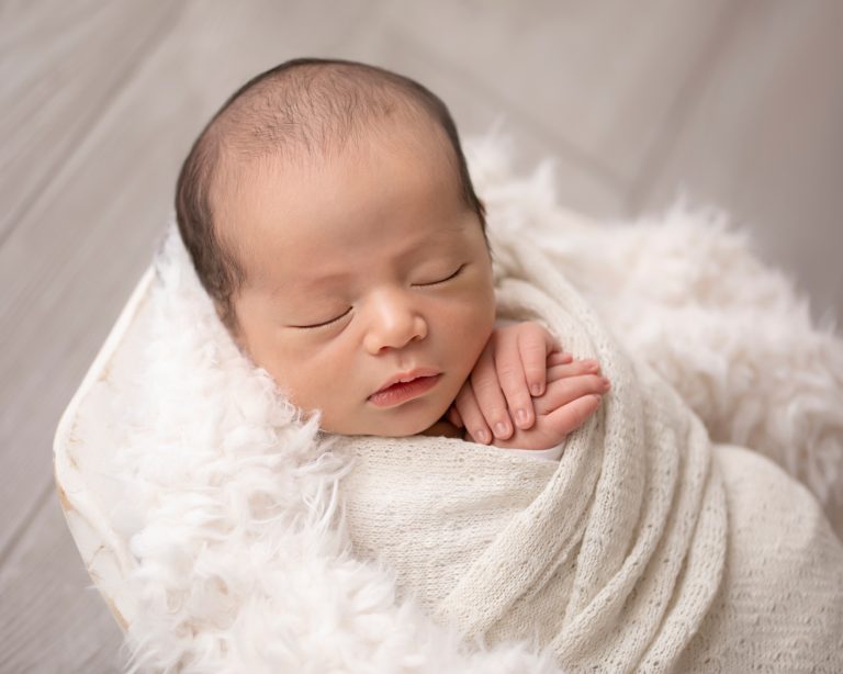 baby close up face and tiny hands tucked under his chin wrapped in white in white fur stuffed bowl against grey floor